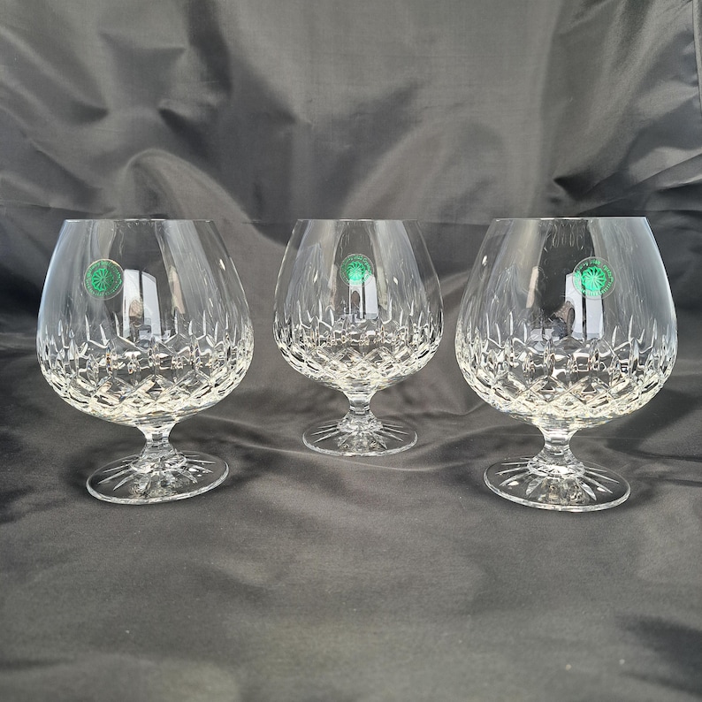 May include: Three clear crystal brandy glasses featuring a deep diamond-cut pattern on the lower bowls and short, sturdy stems with a starburst design on the circular bases. Each glass displays a small, round green sticker on the upper side.