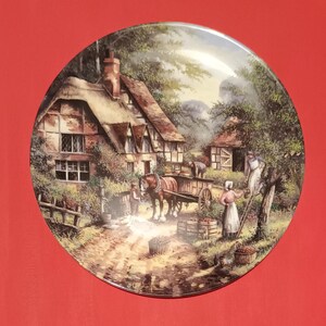 Pode incluir: Um prato de cer&acirc;mica de estilo vintage com uma pintura de uma casa de campo em um ambiente rural. A cena inclui uma carro&ccedil;a puxada por um cavalo, pessoas colhendo ma&ccedil;&atilde;s e uma cerca de madeira.
