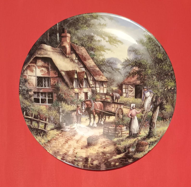 Pode incluir: Um prato de cer&acirc;mica de estilo vintage com uma pintura de uma casa de campo em um ambiente rural. A cena inclui uma carro&ccedil;a puxada por um cavalo, pessoas colhendo ma&ccedil;&atilde;s e uma cerca de madeira.