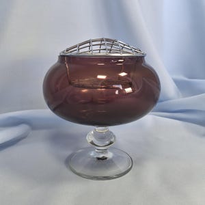 Puede incluir: Un jarr&oacute;n decorativo de cristal color amatista con una base clara y redondeada y una tapa de rejilla de alambre plateado. El jarr&oacute;n tiene un cuerpo bulboso y un tallo esf&eacute;rico transparente. Adecuado para arreglos florales.