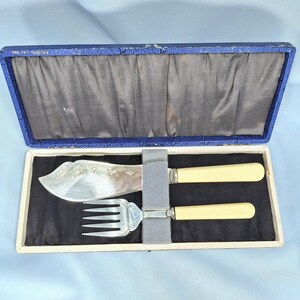 May include: A vintage silver-plated fish serving set, including a knife and fork, presented in a blue and white box. The knife features an ornate floral design on the blade, and both utensils have cream-colored handles.