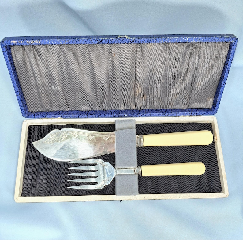 May include: A vintage silver-plated fish serving set, including a knife and fork, presented in a blue and white box. The knife features an ornate floral design on the blade, and both utensils have cream-colored handles.