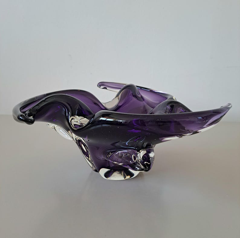 K&ouml;nnte beinhalten: Eine tiefviolette Glasschale mit unregelm&auml;&szlig;igem, gewelltem Rand und einem klaren Glasboden. Die Schale hat eine glatte, gl&auml;nzende Oberfl&auml;che mit durchscheinenden violetten Farbt&ouml;nen, die in klare Glasabschnitte &uuml;bergehen und eine organische Form bilden.