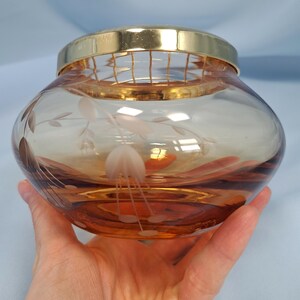 May include: A round, amber-colored glass bowl featuring a delicate etched floral pattern on its surface. The bowl is topped with a removable gold-toned metal lid that includes a built-in wire grid, designed for arranging flowers or holding items.