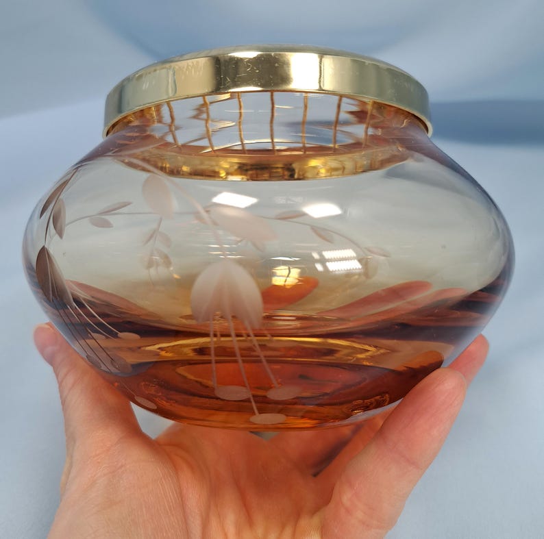May include: A round, amber-colored glass bowl featuring a delicate etched floral pattern on its surface. The bowl is topped with a removable gold-toned metal lid that includes a built-in wire grid, designed for arranging flowers or holding items.