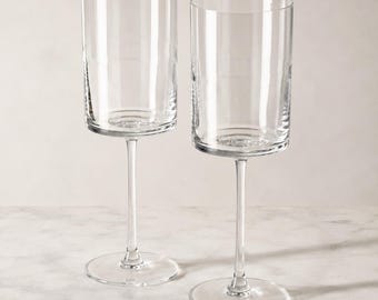May include: Two clear glass vessels with cylindrical bowls and slender stems. Each piece features a flat base and a straight-sided, tall glass body, providing a uniform and transparent appearance for these two matching drinking glasses.