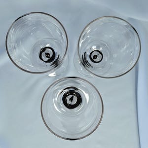 以下が含まれることがあります： 真上から見た、三角形に配置された3つの透明なガラス製ゴブレット。各ゴブレットは滑らかで透明なボウルを備えており、ガラスの底の中心部には特徴的な黒い円形のアクセントが施されています。