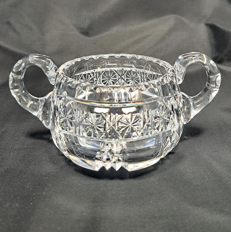 Op de afbeelding: Een suikerpot van helder glas met twee gebogen handvatten en een decoratief geslepen patroon. Het buitenoppervlak vertoont een herhalend sterrenmotief rond het midden, met een geschulpte rand en een gefacetteerde basis voor een reflecterende uitstraling.