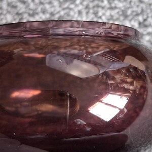 Puede incluir: Primer plano de un cuenco redondo de cristal morado. El cuenco tiene una abertura ancha y un cuerpo redondeado. El cristal refleja la luz, creando reflejos y sombras. La superficie del cuenco tiene peque&ntilde;os ara&ntilde;azos.
