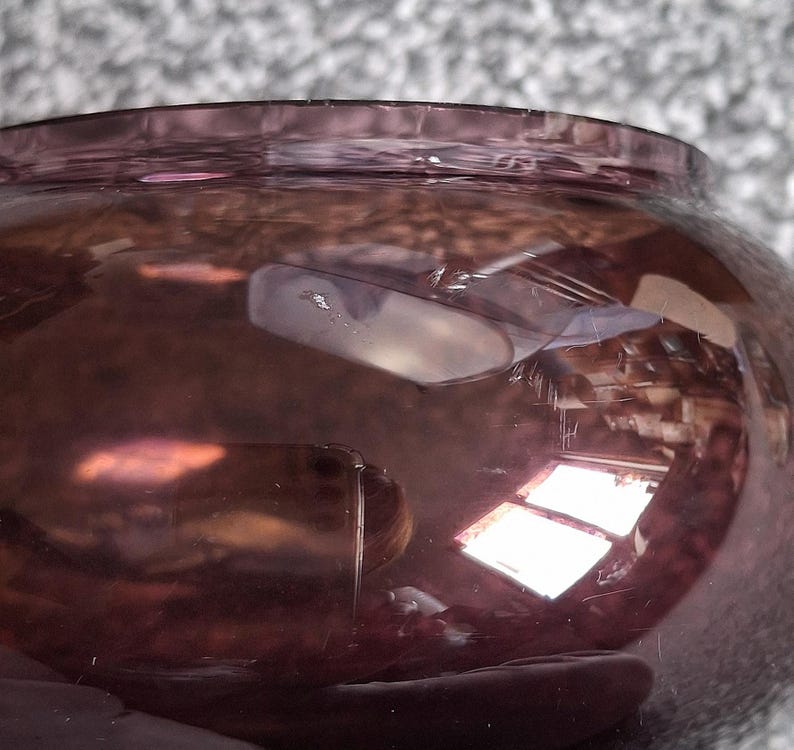 Puede incluir: Primer plano de un cuenco redondo de cristal morado. El cuenco tiene una abertura ancha y un cuerpo redondeado. El cristal refleja la luz, creando reflejos y sombras. La superficie del cuenco tiene peque&ntilde;os ara&ntilde;azos.