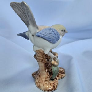 以下が含まれることがあります： 木の枝に止まっている陶器製の鳥の置物。鳥は薄い黄色の頭、青い翼、白い体です。枝は茶色で、緑の葉が付いています。置物は装飾品です。