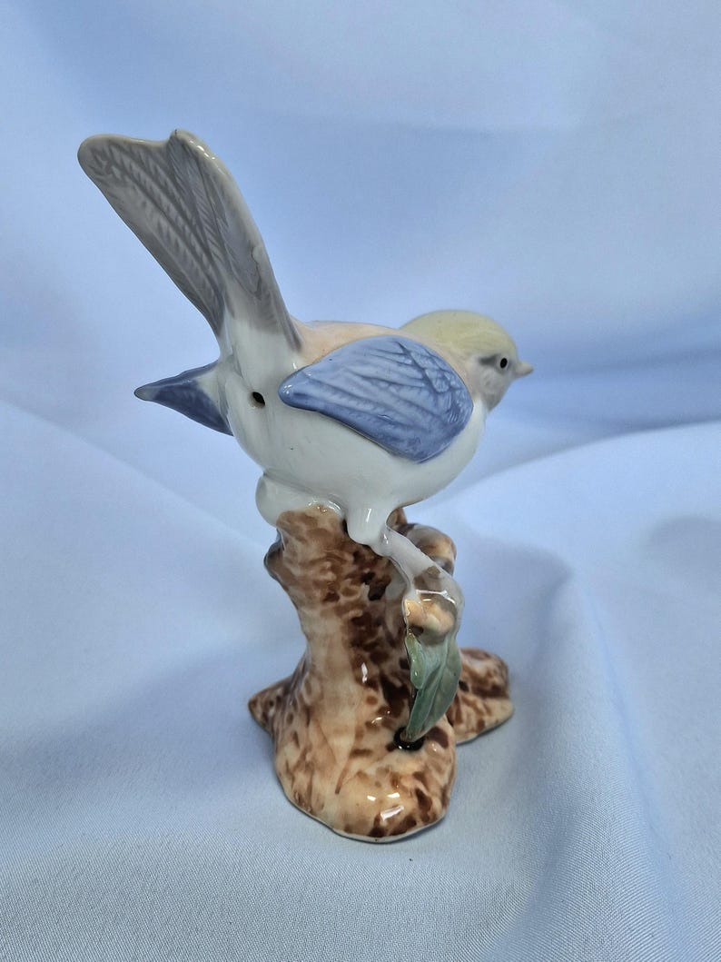 以下が含まれることがあります： 木の枝に止まっている陶器製の鳥の置物。鳥は薄い黄色の頭、青い翼、白い体です。枝は茶色で、緑の葉が付いています。置物は装飾品です。