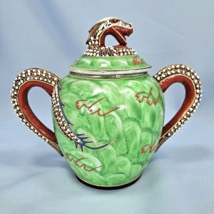 Pu&ograve; includere: Una zuccheriera in ceramica verde con coperchio a testa di drago. La zuccheriera ha due manici con un design a perline rosse e bianche. Il corpo della zuccheriera presenta un motivo a vortice verde con accenti rossi e blu.
