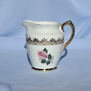 May include: A cream-coloured porcelain creamer with a gold handle and trim. The creamer features a pink rose with green leaves and a decorative black and gold band. The base is also trimmed in gold.