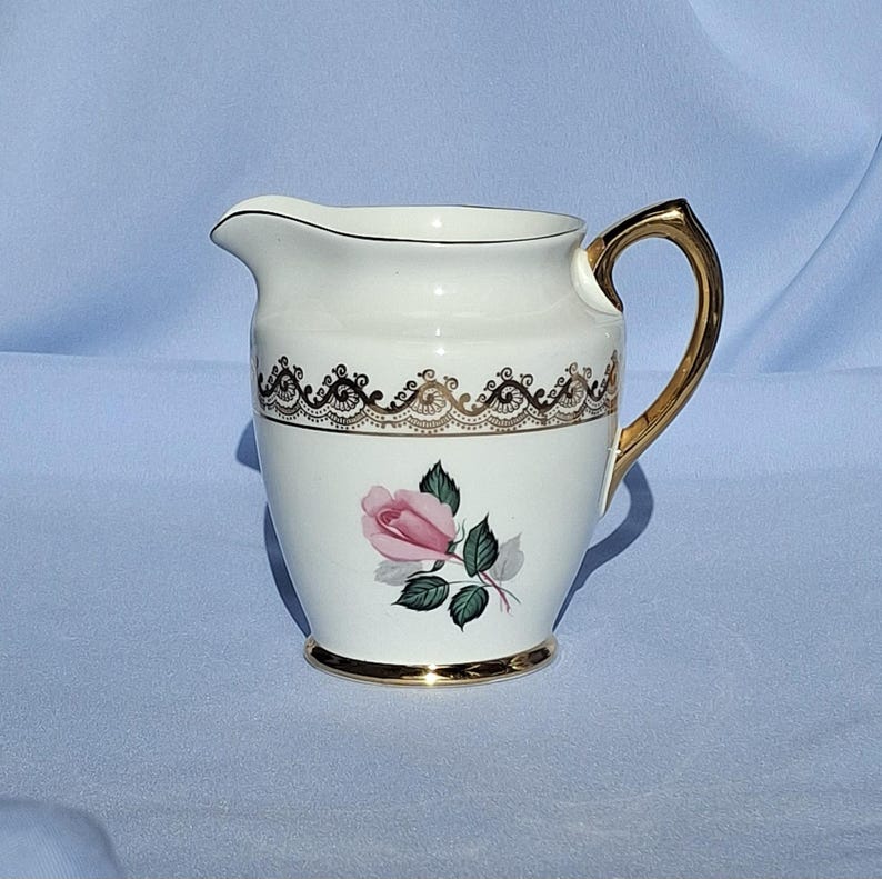 May include: A cream-coloured porcelain creamer with a gold handle and trim. The creamer features a pink rose with green leaves and a decorative black and gold band. The base is also trimmed in gold.