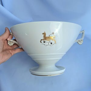 May include: White ceramic serving bowl with gold deer and floral design. The bowl has two handles with gold accents and a pedestal base. The deer is gold with black details. The bowl is likely for serving food or as a decorative piece.