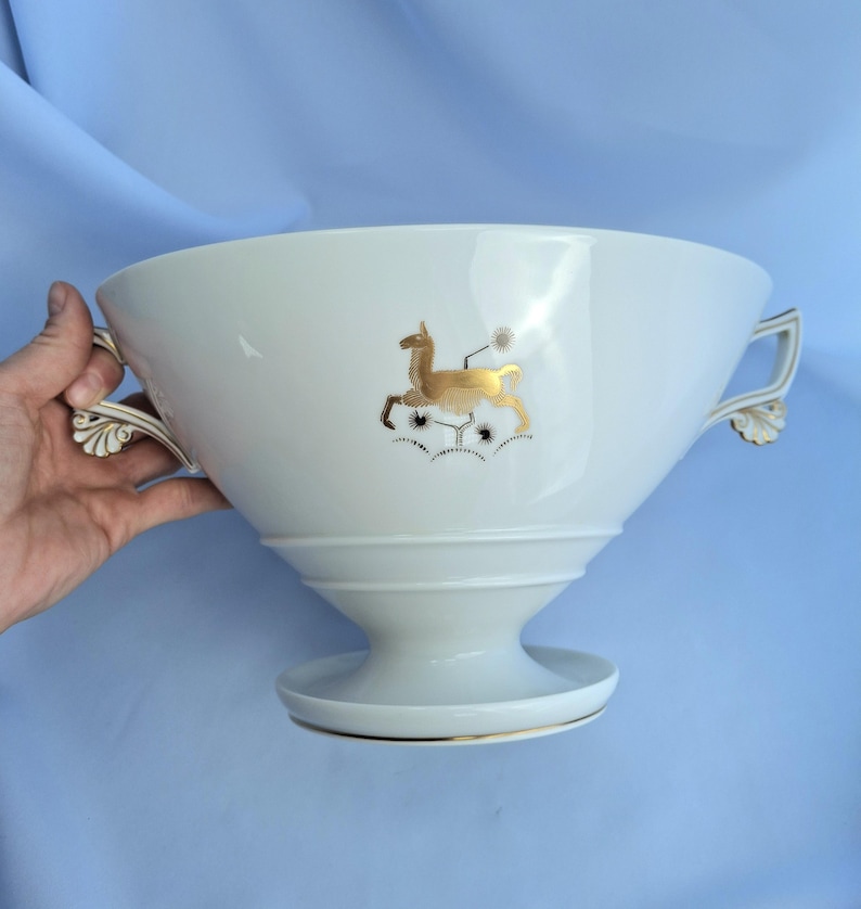 May include: White ceramic serving bowl with gold deer and floral design. The bowl has two handles with gold accents and a pedestal base. The deer is gold with black details. The bowl is likely for serving food or as a decorative piece.
