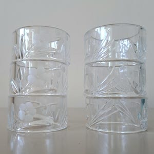 May include: A set of six clear glass napkin rings arranged in two stacks of three. Each ring features a delicate etched floral design with frosted petals and leaves, providing a subtle texture against the smooth, transparent glass surface.