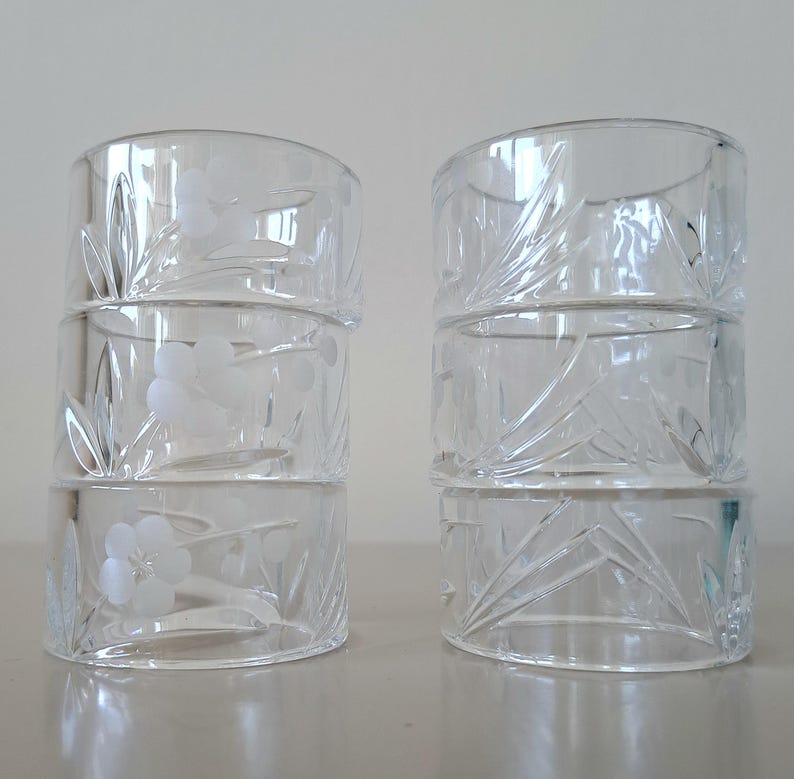 May include: A set of six clear glass napkin rings arranged in two stacks of three. Each ring features a delicate etched floral design with frosted petals and leaves, providing a subtle texture against the smooth, transparent glass surface.