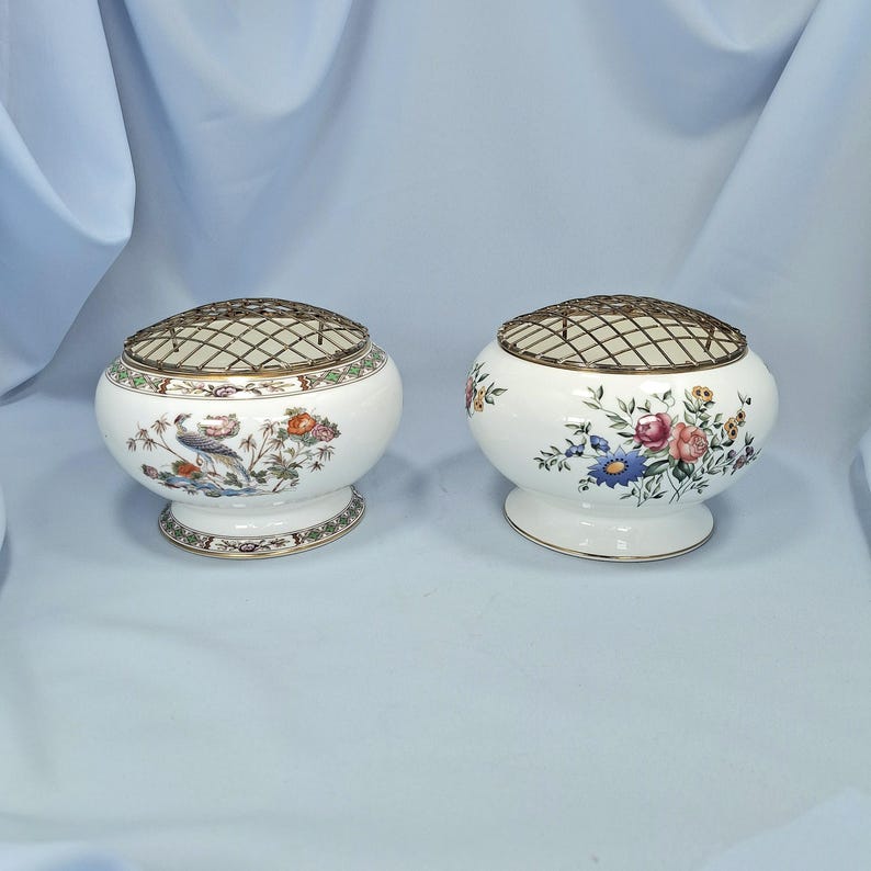 Puede incluir: Dos floreros de cer&aacute;mica blanca con tapas de rejilla met&aacute;lica. Uno presenta un dise&ntilde;o de p&aacute;jaros y flores, el otro tiene un estampado floral. Ambos tienen una base de pedestal y son adecuados para arreglos florales.
