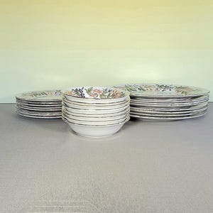 以下が含まれることがあります： 花柄とベリー柄の白いセラミック食器セット。 金色の縁が付いたお皿とボウルのスタックが含まれています。 お皿とボウルは、明るい灰色の表面に配置されています。