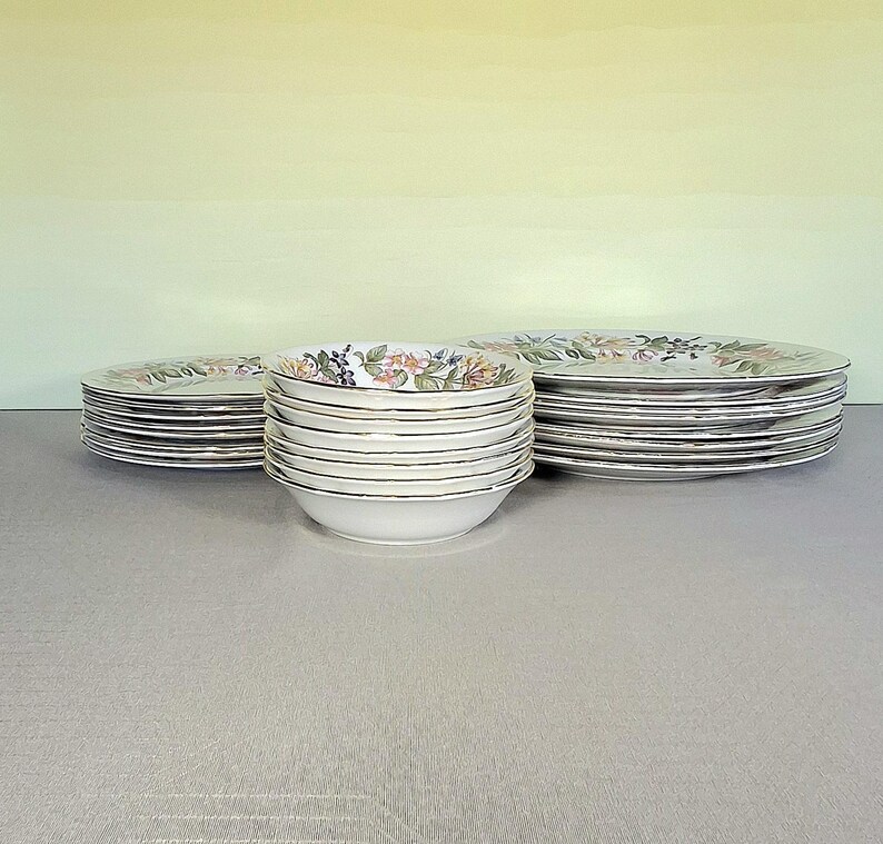 以下が含まれることがあります： 花柄とベリー柄の白いセラミック食器セット。 金色の縁が付いたお皿とボウルのスタックが含まれています。 お皿とボウルは、明るい灰色の表面に配置されています。