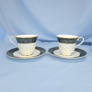May include: Two white porcelain teacups and matching saucers featuring a dark blue band with a gold floral pattern. Each cup has a curved handle and a gold rim, while the saucers display a coordinating dark blue and gold floral border design.
