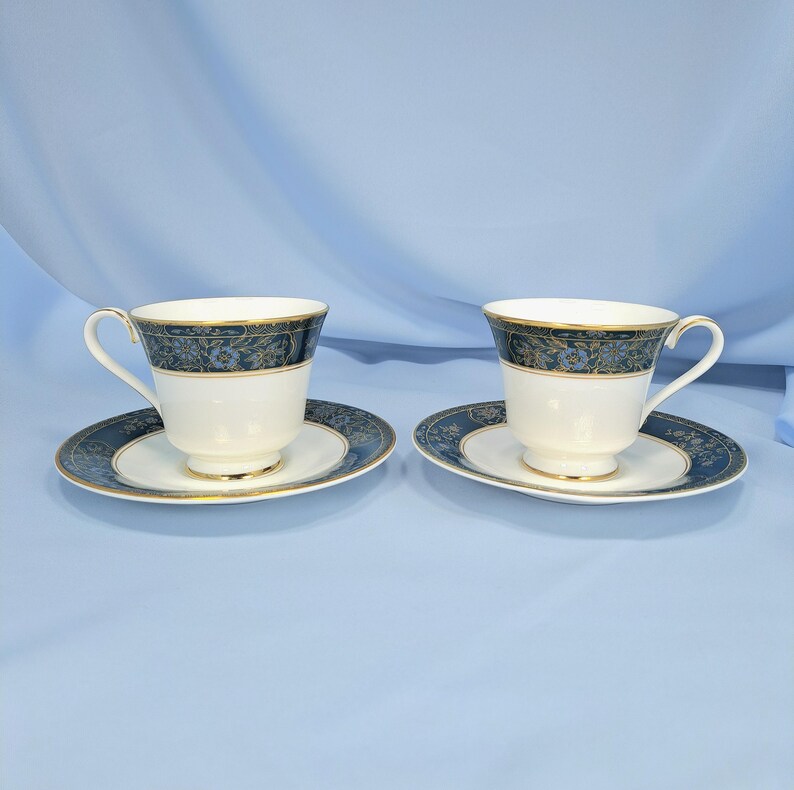 May include: Two white porcelain teacups and matching saucers featuring a dark blue band with a gold floral pattern. Each cup has a curved handle and a gold rim, while the saucers display a coordinating dark blue and gold floral border design.