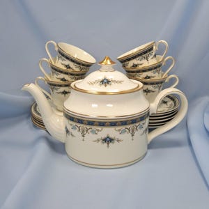 May include: A porcelain tea set featuring a teapot with a lid and multiple matching teacups and saucers. The pieces are cream-coloured with a decorative border of blue, gold, and light blue floral patterns, finished with gold trim on the edges.