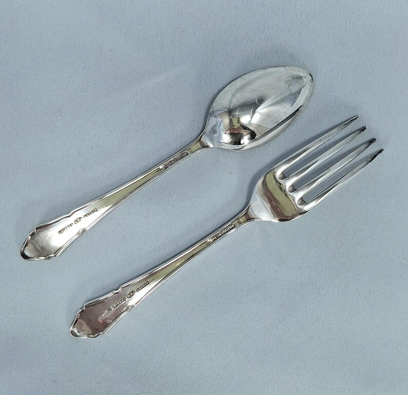 May include: A silver spoon and fork set. The spoon has an oval bowl and a long handle. The fork has four tines and a long handle. Both pieces have a decorative edge and are set against a light blue background.