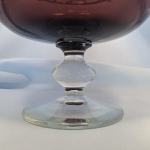 Puede incluir: Una copa de cristal transparente con un cuenco ancho, profundo y morado. La copa tiene un tallo claro y bulboso y una base circular. El cuenco es de color morado oscuro, y el tallo y la base son transparentes.