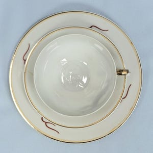 May include: A white teacup, saucer, and plate set with gold trim and maroon accent designs. The teacup is centered on the saucer, which sits on the plate. The set is on a light blue surface.