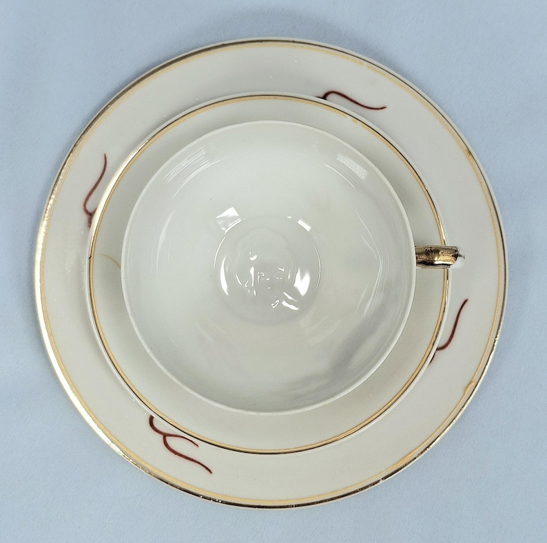 May include: A white teacup, saucer, and plate set with gold trim and maroon accent designs. The teacup is centered on the saucer, which sits on the plate. The set is on a light blue surface.