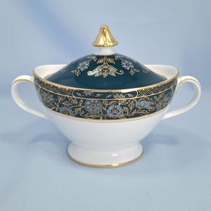 May include: A white porcelain sugar bowl with two handles and a matching lid. The lid and upper rim feature a dark teal band decorated with intricate gold filigree and light blue floral patterns. The lid is topped with a gold-colored finial.