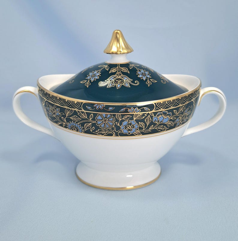 May include: A white porcelain sugar bowl with two handles and a matching lid. The lid and upper rim feature a dark teal band decorated with intricate gold filigree and light blue floral patterns. The lid is topped with a gold-colored finial.