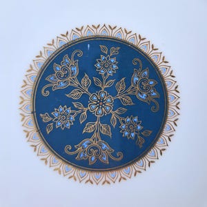 Op de afbeelding: Een rond keramieken bord met een diepblauw middenstuk versierd met gouden en lichtblauwe bloemmotieven. Het ontwerp bevat een centrale bloem omringd door vier kleinere bloesems en bladeren, omlijst door een gouden en blauwe geschulpte rand.