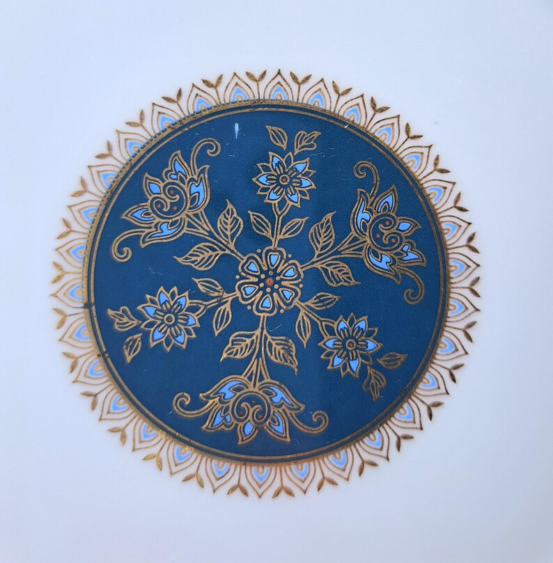 Op de afbeelding: Een rond keramieken bord met een diepblauw middenstuk versierd met gouden en lichtblauwe bloemmotieven. Het ontwerp bevat een centrale bloem omringd door vier kleinere bloesems en bladeren, omlijst door een gouden en blauwe geschulpte rand.