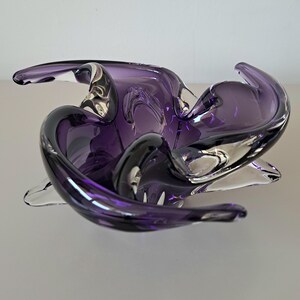 K&ouml;nnte beinhalten: Eine tiefviolette Glasschale mit organischer, freier Form, die wirbelnde, durchscheinende Schichten und dicke, geschwungene Kanten aufweist. Die Schale hat eine polierte, reflektierende Oberfl&auml;che mit unterschiedlich intensiven Violettt&ouml;nen im Glas.