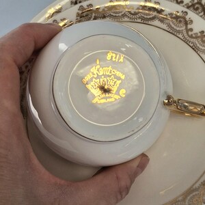 May include: A white Bone China teacup and saucer set with gold detailing. The teacup base displays "Bone Kentchina" and a crown logo. The saucer has a gold filigree pattern around the rim. A classic tea set.