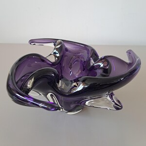K&ouml;nnte beinhalten: Eine tiefviolette Glasschale mit organischem, freiem Design und wellenf&ouml;rmigen R&auml;ndern. Das durchscheinende Material geht von dunklen Violettt&ouml;nen zu klarem Glas am Boden &uuml;ber und erzeugt so ein flie&szlig;endes, skulpturales Erscheinungsbild.