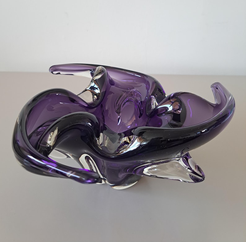 K&ouml;nnte beinhalten: Eine tiefviolette Glasschale mit organischem, freiem Design und wellenf&ouml;rmigen R&auml;ndern. Das durchscheinende Material geht von dunklen Violettt&ouml;nen zu klarem Glas am Boden &uuml;ber und erzeugt so ein flie&szlig;endes, skulpturales Erscheinungsbild.