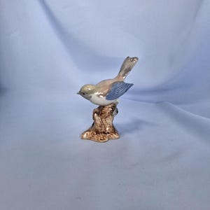 以下が含まれることがあります： 茶色の切り株に止まっている陶器製の鳥の置物。鳥は薄茶色の体、青い翼、灰色の尾を持っています。この置物は細部までこだわっており、手描きで繊細なデザインが施されているようです。