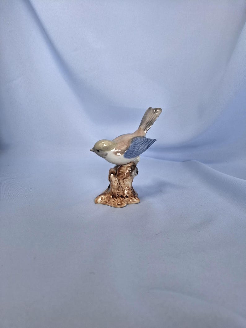 以下が含まれることがあります： 茶色の切り株に止まっている陶器製の鳥の置物。鳥は薄茶色の体、青い翼、灰色の尾を持っています。この置物は細部までこだわっており、手描きで繊細なデザインが施されているようです。