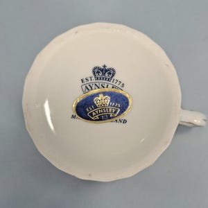 Peut inclure: Une tasse &agrave; th&eacute; blanche avec un bord festonn&eacute;, portant une &eacute;tiquette bleue et dor&eacute;e Aynsley England. L'&eacute;tiquette comprend le texte "EST. 1775" et un motif de couronne. La tasse a une petite anse et est pos&eacute;e sur un fond bleu clair.