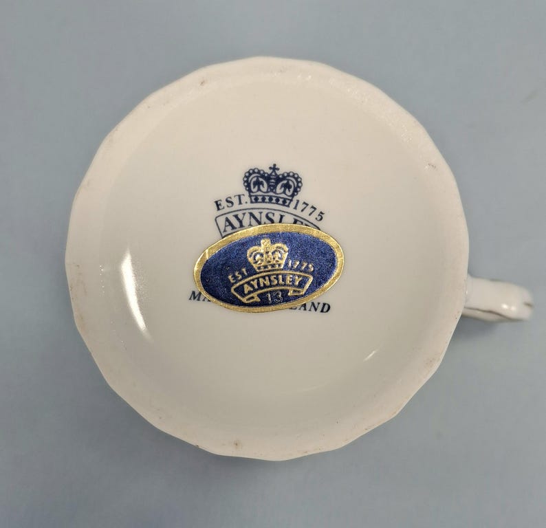 Peut inclure: Une tasse &agrave; th&eacute; blanche avec un bord festonn&eacute;, portant une &eacute;tiquette bleue et dor&eacute;e Aynsley England. L'&eacute;tiquette comprend le texte "EST. 1775" et un motif de couronne. La tasse a une petite anse et est pos&eacute;e sur un fond bleu clair.
