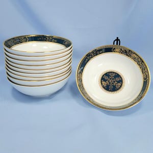 Op de afbeelding: Een set van tien witte keramische kommen met een donkerblauwe rand met een gouden bloemmotief en een bijpassend cirkelvormig goud en blauw bloemmotief in het midden van elke kom. De kommen zijn gestapeld met &eacute;&eacute;n kom apart weergegeven.