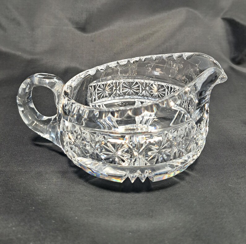 Op de afbeelding: Een creamer van helder glas met een gedetailleerd ster- en kruispatroon. De kan heeft een gebogen handvat aan &eacute;&eacute;n kant, een geschulpte rand en een kleine schenktuit, wat zorgt voor een reflecterend en gefacetteerd oppervlak over het geheel.
