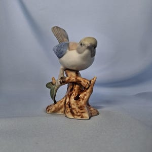 以下が含まれることがあります： 茶色の切り株に止まっている陶器の鳥の置物。鳥は白い体、薄緑色の頭、青と茶色の翼をしています。緑の葉が木の枝に付いています。背景は水色です。