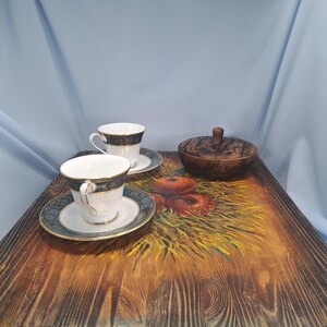 May include: A rectangular wooden serving tray with a painted design of red apples on a bed of yellow straw. Two white porcelain teacups with blue and gold patterned rims and saucers sit on the tray alongside a small, round, dark wooden lidded bowl.