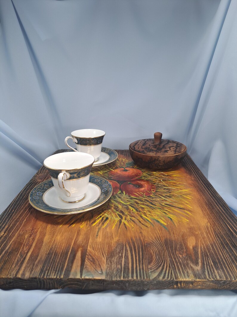 May include: A rectangular wooden serving tray with a painted design of red apples on a bed of yellow straw. Two white porcelain teacups with blue and gold patterned rims and saucers sit on the tray alongside a small, round, dark wooden lidded bowl.